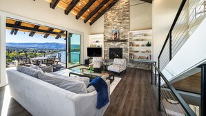 A spacious living area with a large sofa, two armchairs, a coffee table, and a fireplace. There is a balcony with a view of the mountains and a pool.