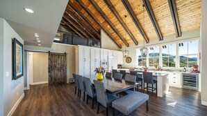 A spacious dining area with a wooden table and chairs, a kitchen with large windows, and a loft space with a wooden ceiling.