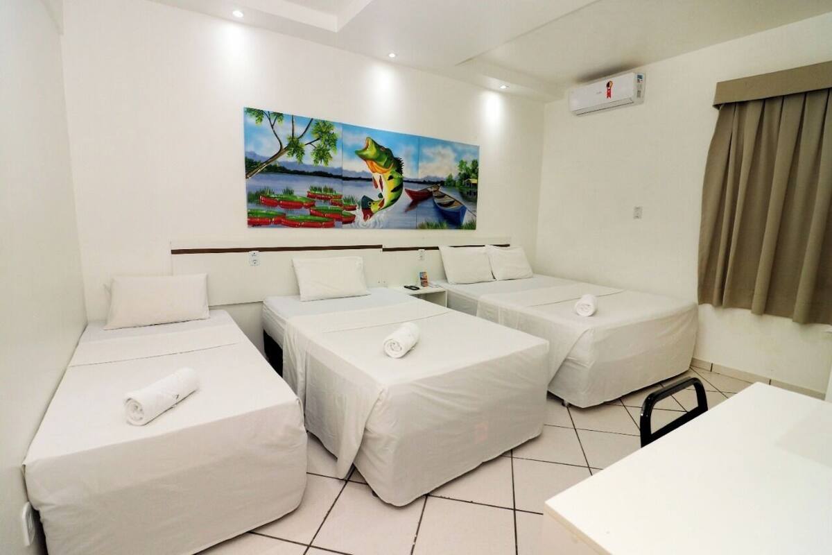 A hotel room with three single beds, a small table, and a painting of a tropical scene on the wall.