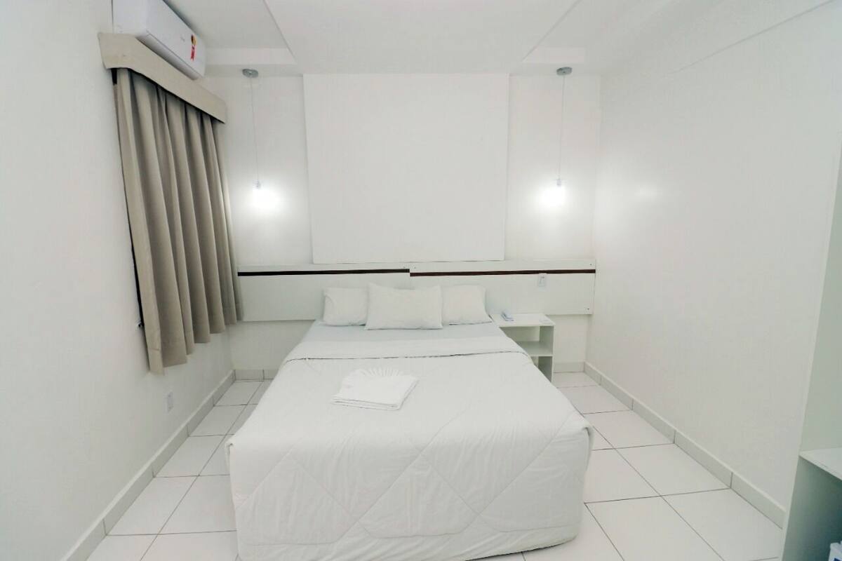 A clean, white hotel room with a bed, a nightstand, and a wall-mounted air conditioner.