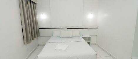 Una habitación de hotel limpia y blanca con una cama, una mesita de noche y un aire acondicionado de pared.