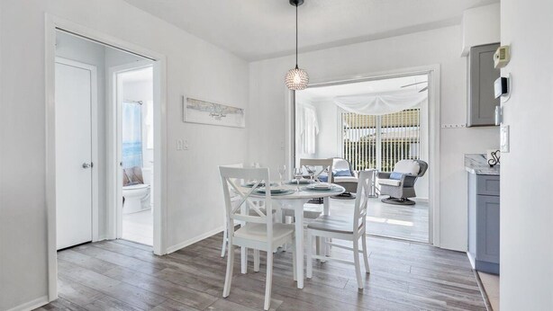 A bright, open-plan living area with a dining table set for four, a kitchen area with a blue cabinet, and a view through a glass door to an outdoor balcony.