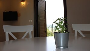 A room with a table, two white chairs, a potted plant, a TV on the wall, and a door leading to a balcony with a view.