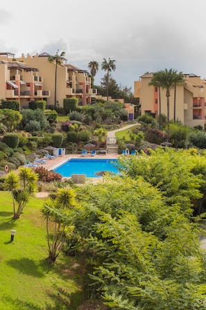 A pool area with lounge chairs and a clear blue pool surrounded by lush greenery and multi-story buildings.