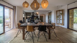 Un vaste espace dédié à la salle à manger, avec une grande table et des chaises, une cuisine à l’arrière-plan et de grandes fenêtres offrant une vue sur l’extérieur.