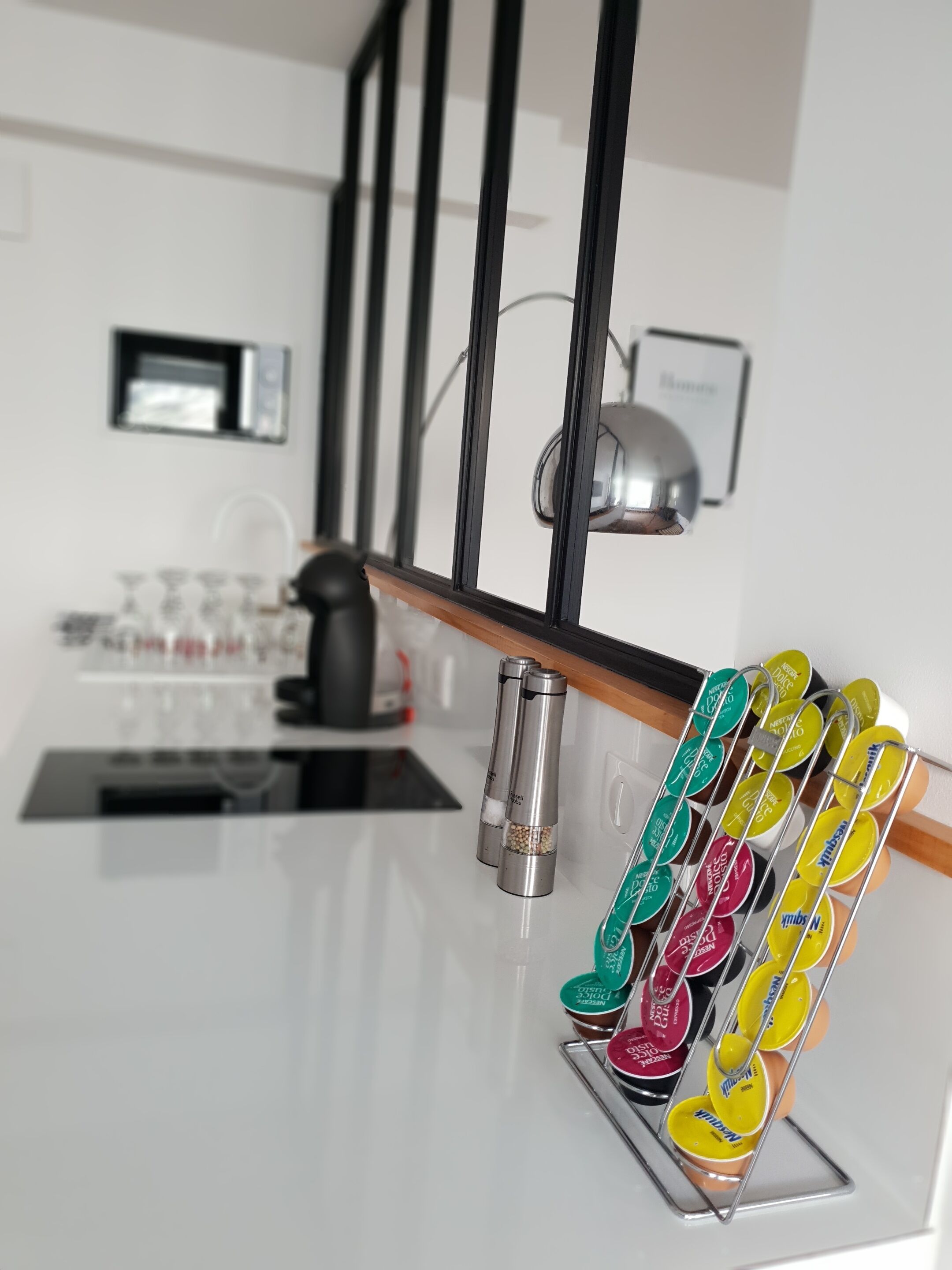 Een keuken met een wit aanrecht, een raam met zwarte omlijsting en een metalen rek met kleurrijke kruidenpotjes.