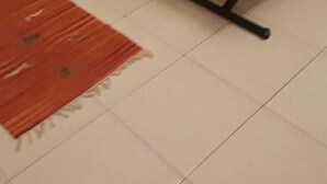 Uma sala de estar com duas cadeiras de madeira, uma mesa de centro de vidro com uma revista, uma televisão pequena e um tapete vermelho.