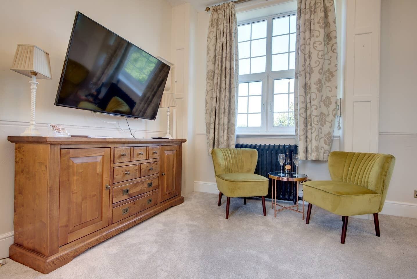 A living room with a wooden cabinet, a flat-screen TV, two green armchairs, a small round table with a candle and glasses, and a window with patterned curtains.
