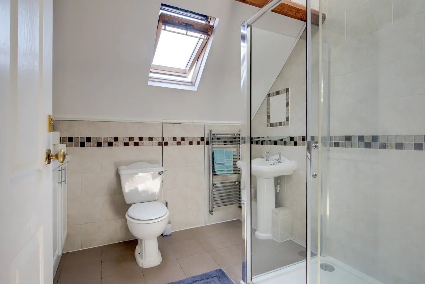 Een moderne badkamer met toilet, wastafel en douche. Boven het toilet bevindt zich een dakraam en de douche is omgeven door glas.