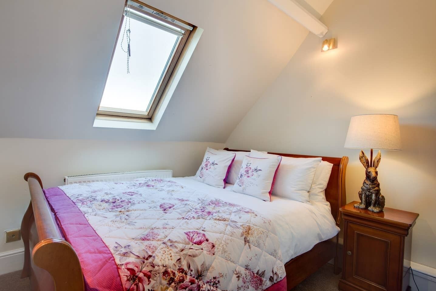 Une chambre avec un plafond en pente, un puits de lumière, un lit avec une literie à motifs floraux, une table de chevet avec une lampe et un banc en bois.