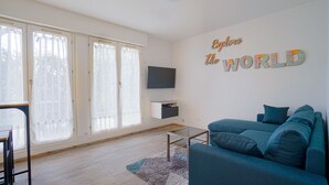 Un salon moderne avec un canapé bleu, un téléviseur à écran plat fixé au mur et une grande fenêtre ornée de rideaux légers.