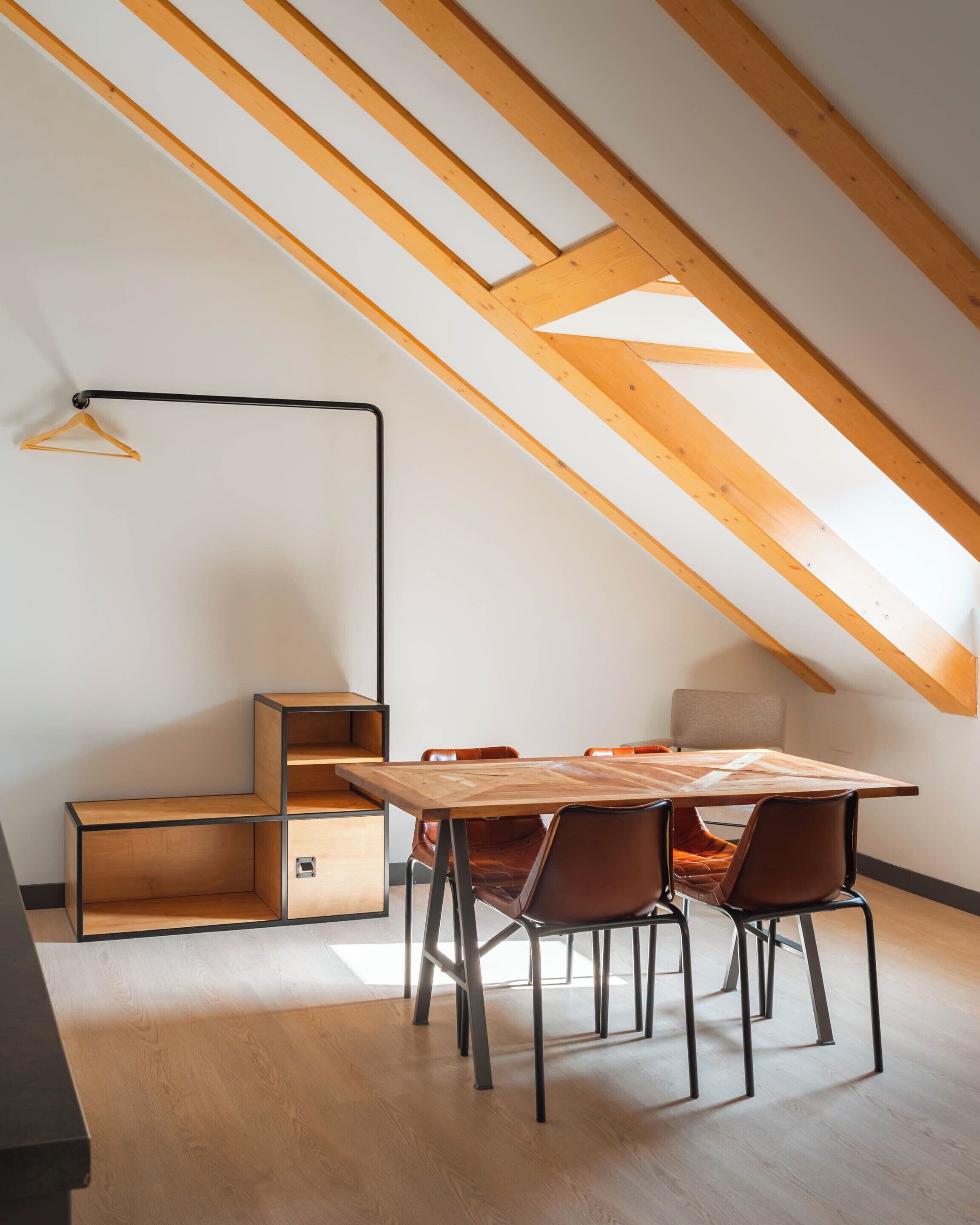 A modern loft bedroom with a sloped ceiling, exposed wooden beams, a wooden dining table with chairs, a hanging light fixture, and built-in storage units.