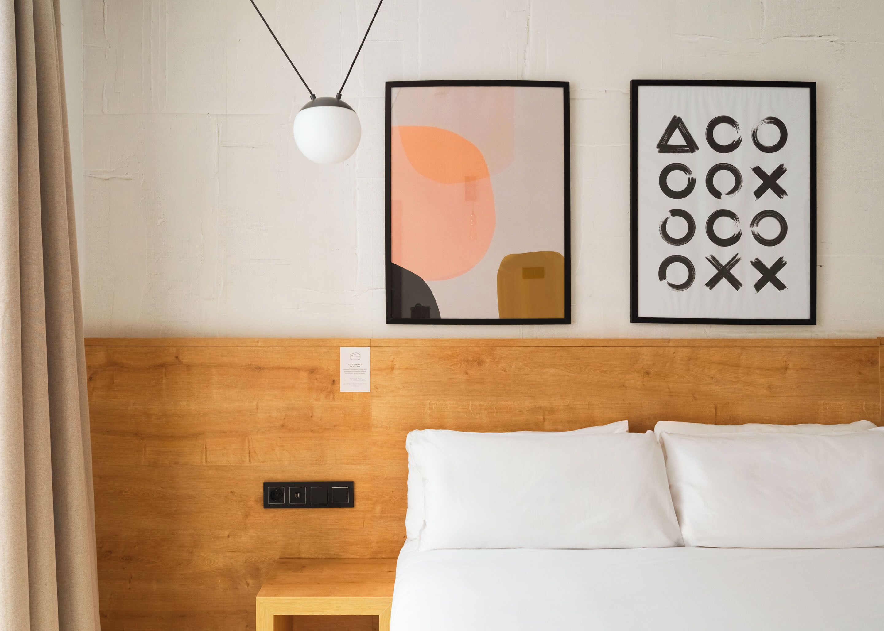 A hotel room with a wooden headboard, a bed with white linens, a bedside table, and two framed artworks on the wall.