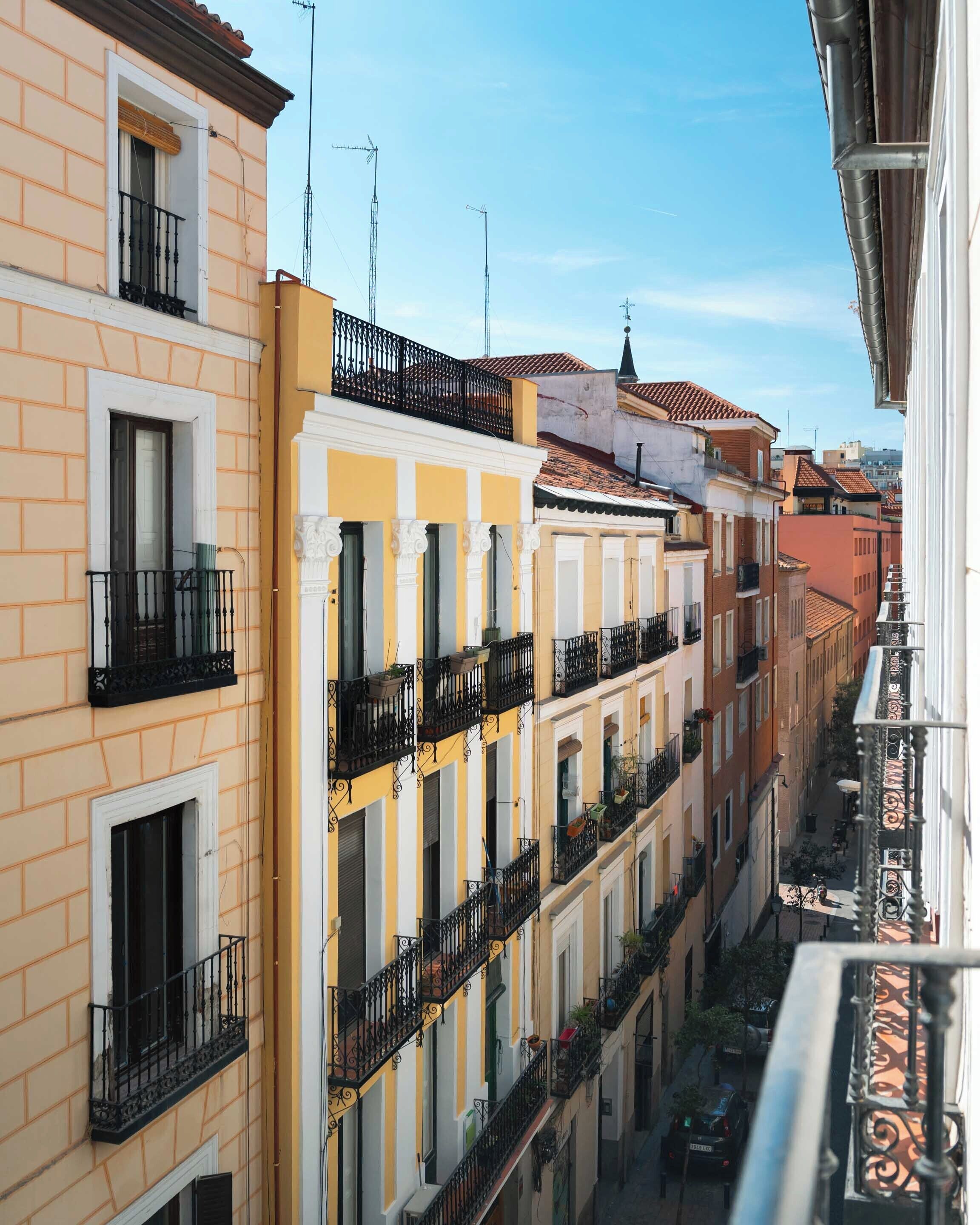 A row of multi-story buildings with balconies, a narrow street, and a clear sky.