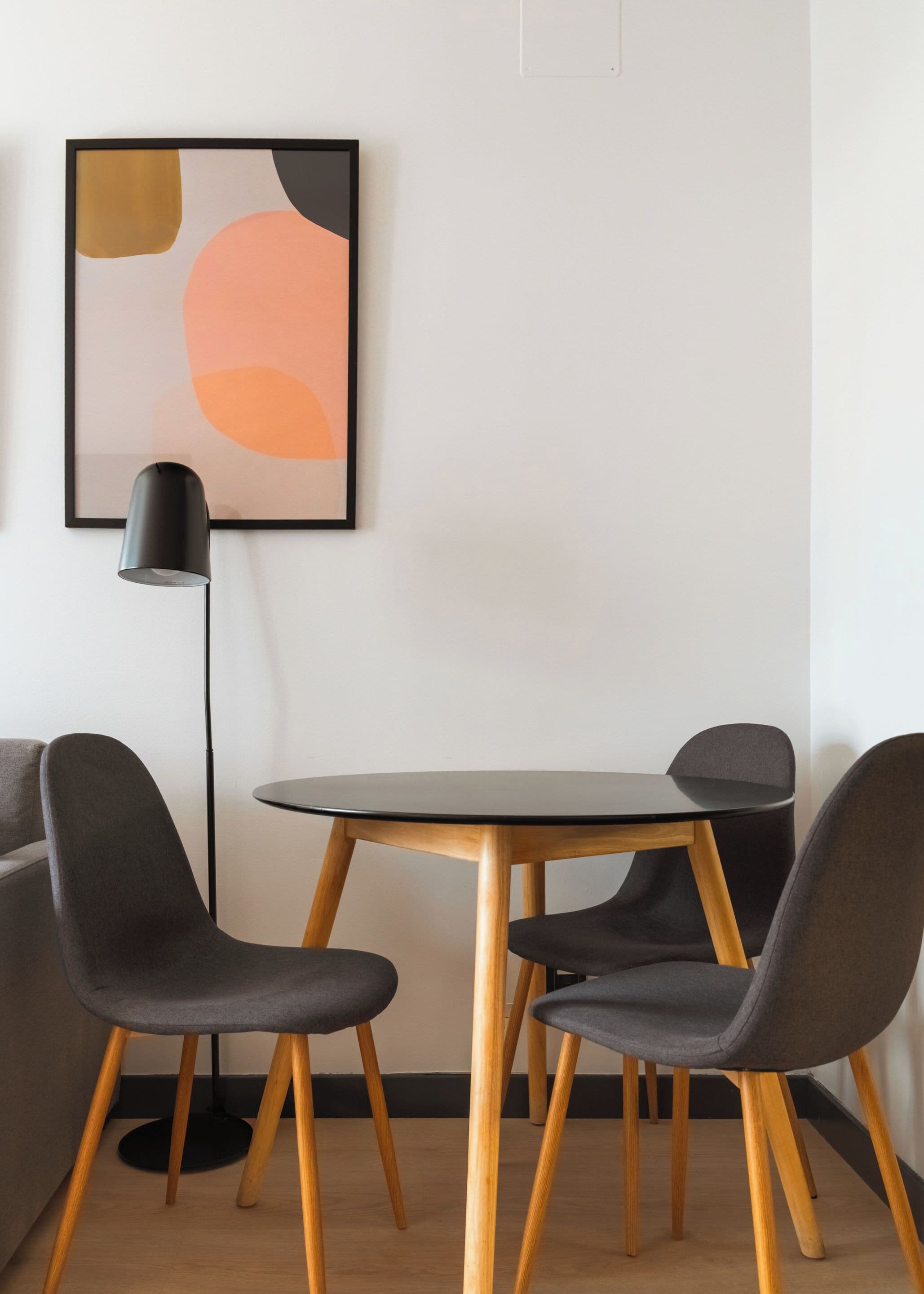 A modern room with a round wooden table, two grey chairs, a floor lamp, and a minimalist abstract painting on the wall.