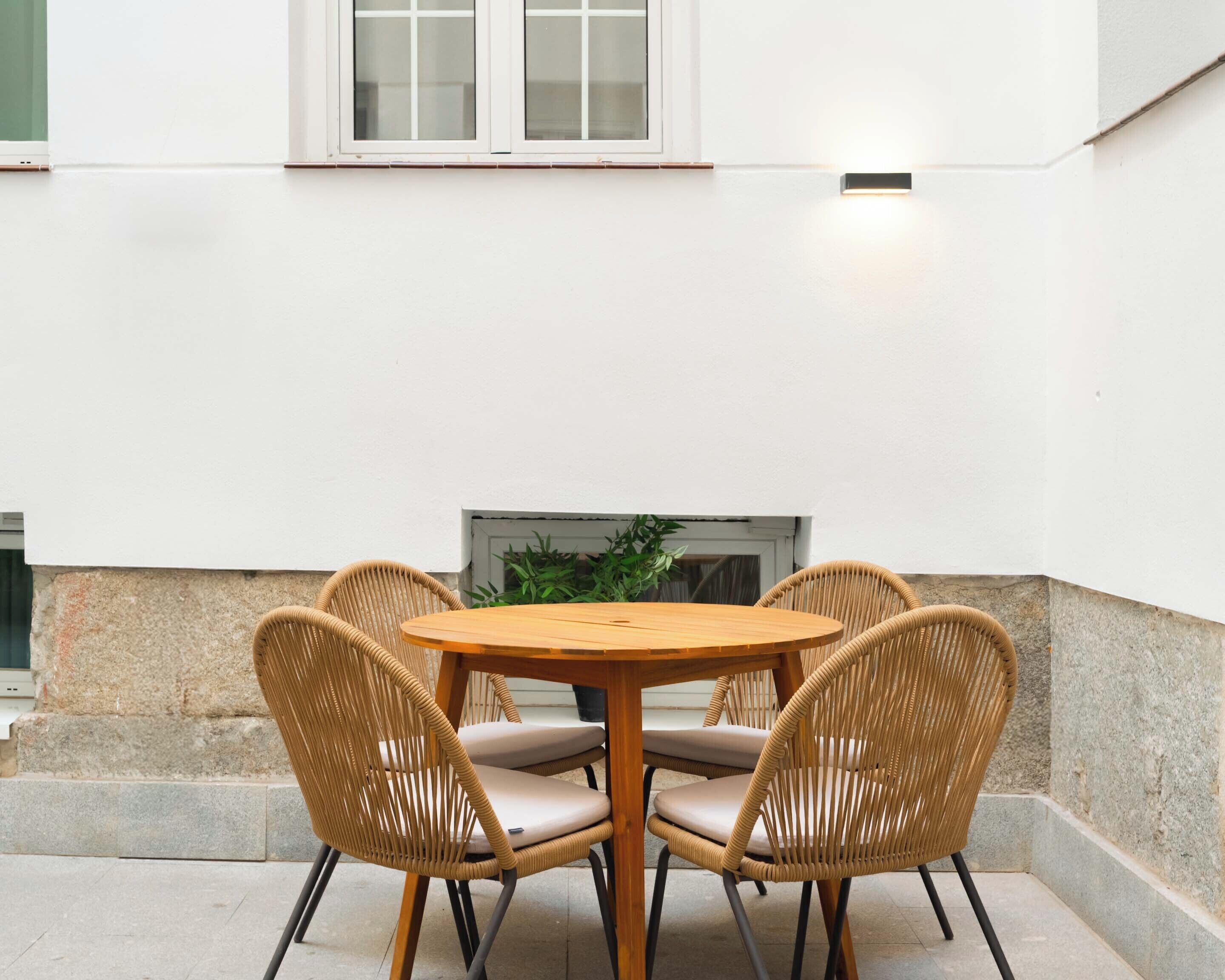A round wooden table with four wicker chairs, a white wall, and a small window above.