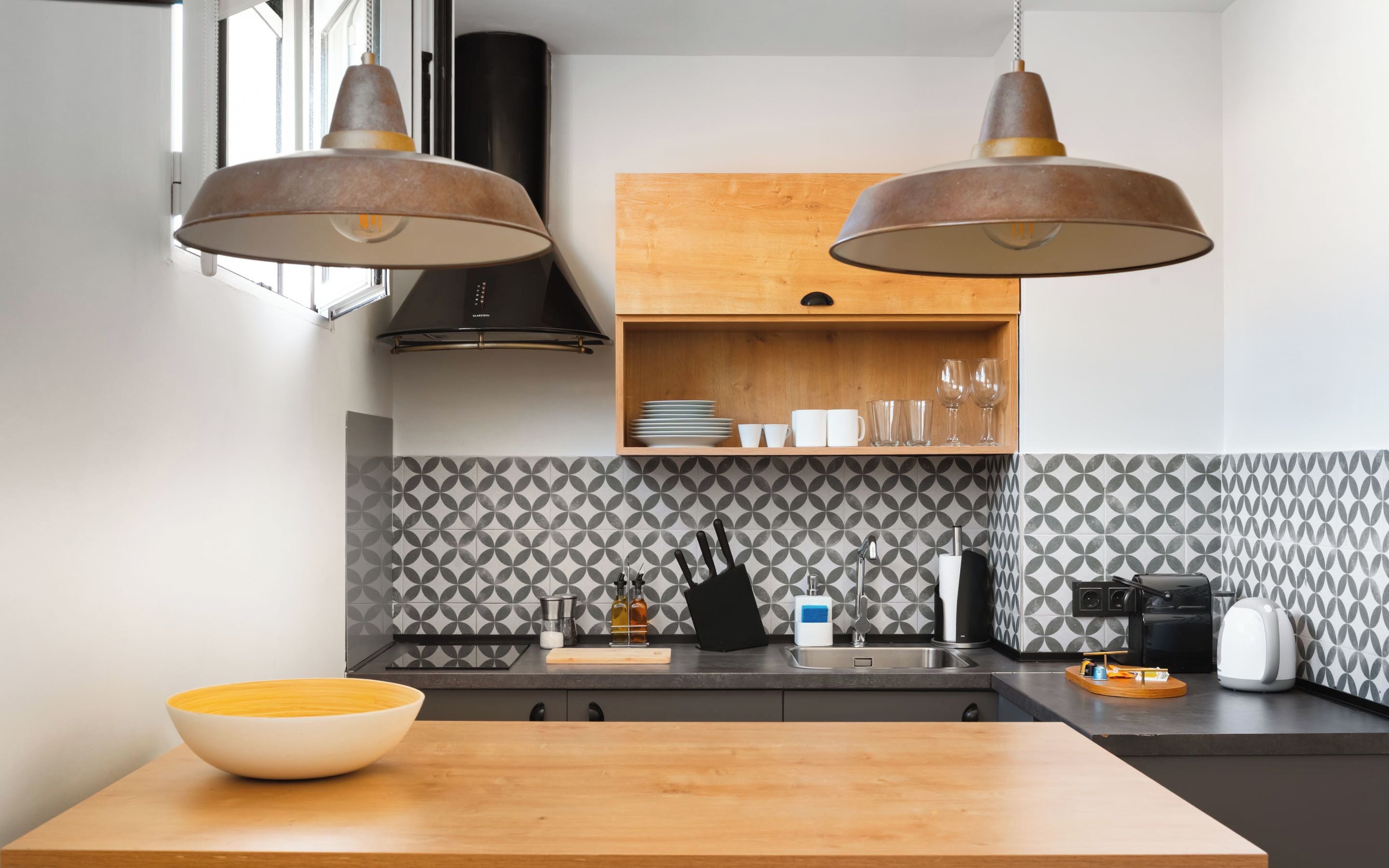A modern kitchen with two pendant lights, a wooden countertop, a sink, and a backsplash with a geometric pattern.