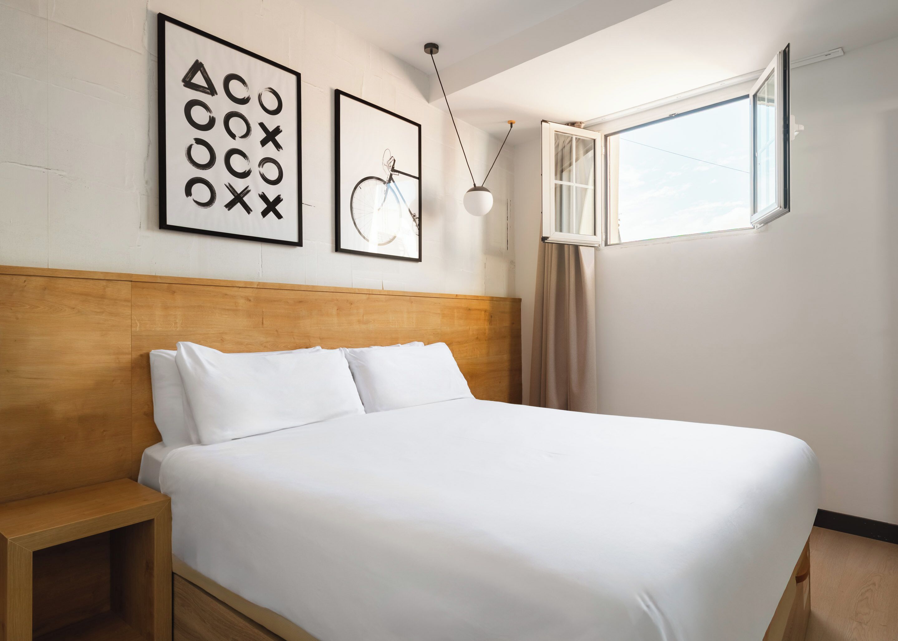 Una habitación de hotel con cabecera de madera, una cama grande con ropa blanca, una mesita de noche, una lámpara de techo y una ventana con cortinas.