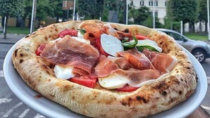 Pizza con jamón serrano y mozzarella en un plato, con una iglesia y palmeras al fondo.