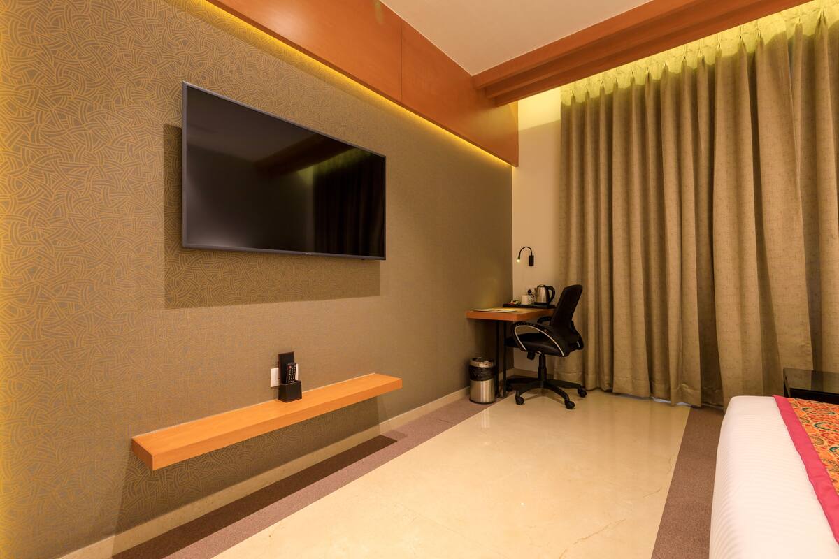 A modern hotel room with a flat-screen TV mounted on the wall, a desk with a chair, a small trash bin, and a bed with a patterned bedspread.