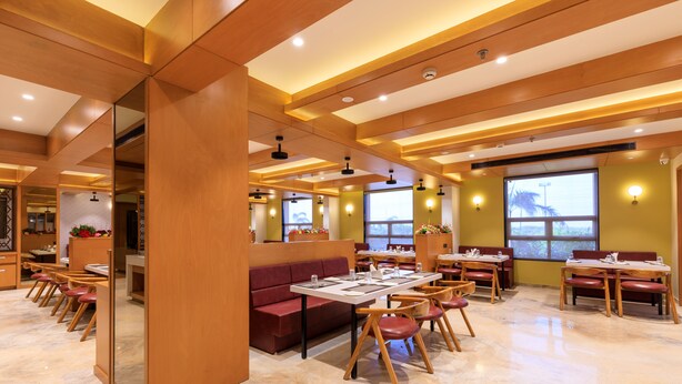 A modern restaurant interior with wooden ceiling beams, marble flooring, and red seating. Tables are set with white tablecloths and red chairs. - Lords Eco Inn Jamnagar (Jamnagar)