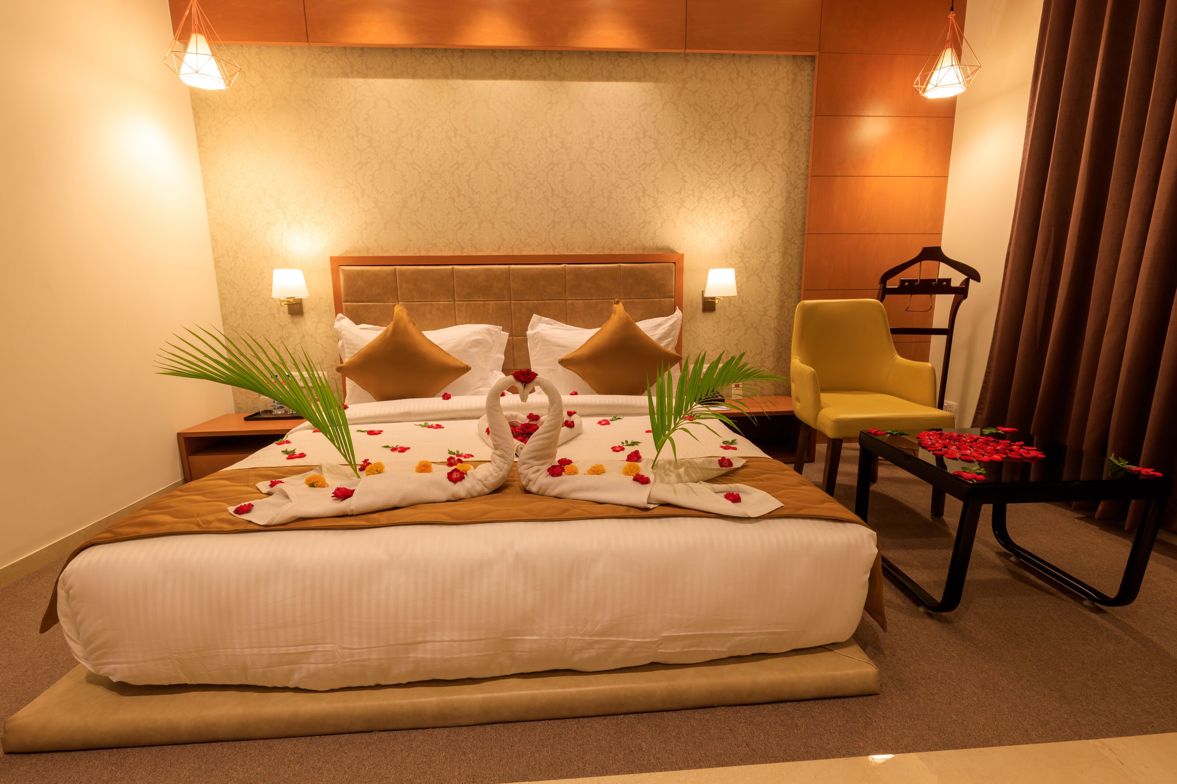 Camera d'albergo con un letto grande, due lampade a parete, una testiera in legno, un comodino con lampada, una sedia e un tavolino con fiori rossi.