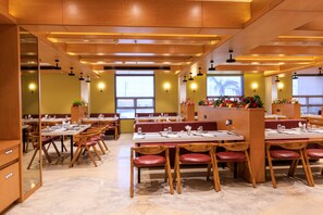 A restaurant with wooden tables and chairs, large windows, and wall-mounted lights. - Lords Eco Inn Jamnagar (Jamnagar)