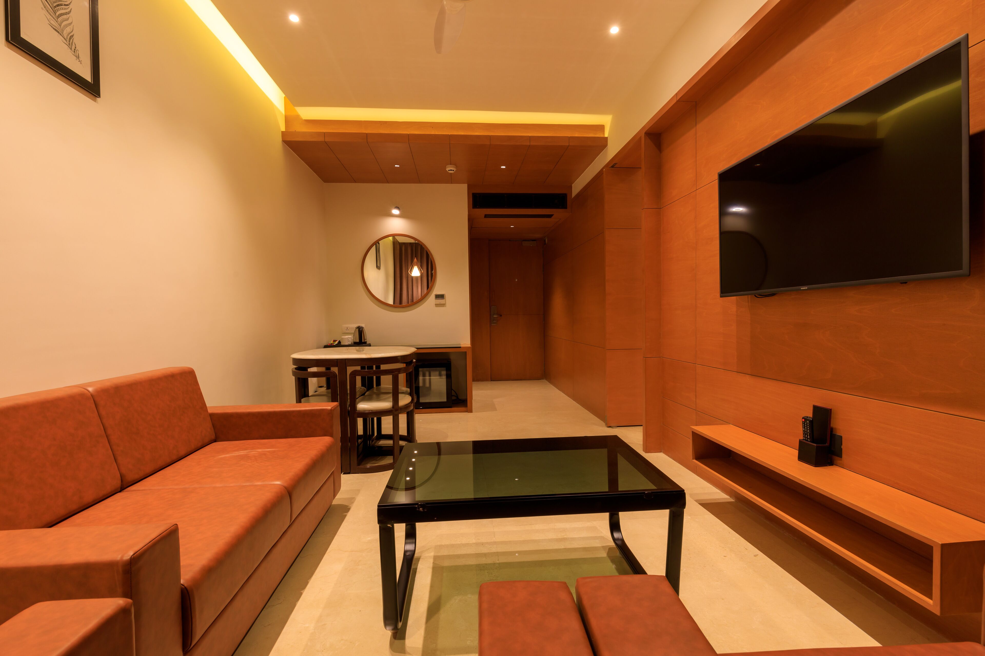 A modern living room with a sofa, a glass-top coffee table, a TV mounted on the wall, and a dining area in the background.