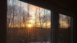 Näkymä ikkunasta lumiseen maisemaan, jossa on paljaita puita ja auringonlasku.