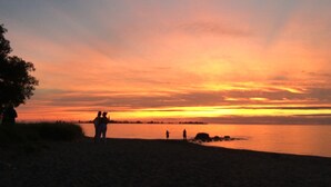 Auringonlasku tyynen vesistön yllä, ja rannalla näkyy ihmisten hahmoja siluettina.