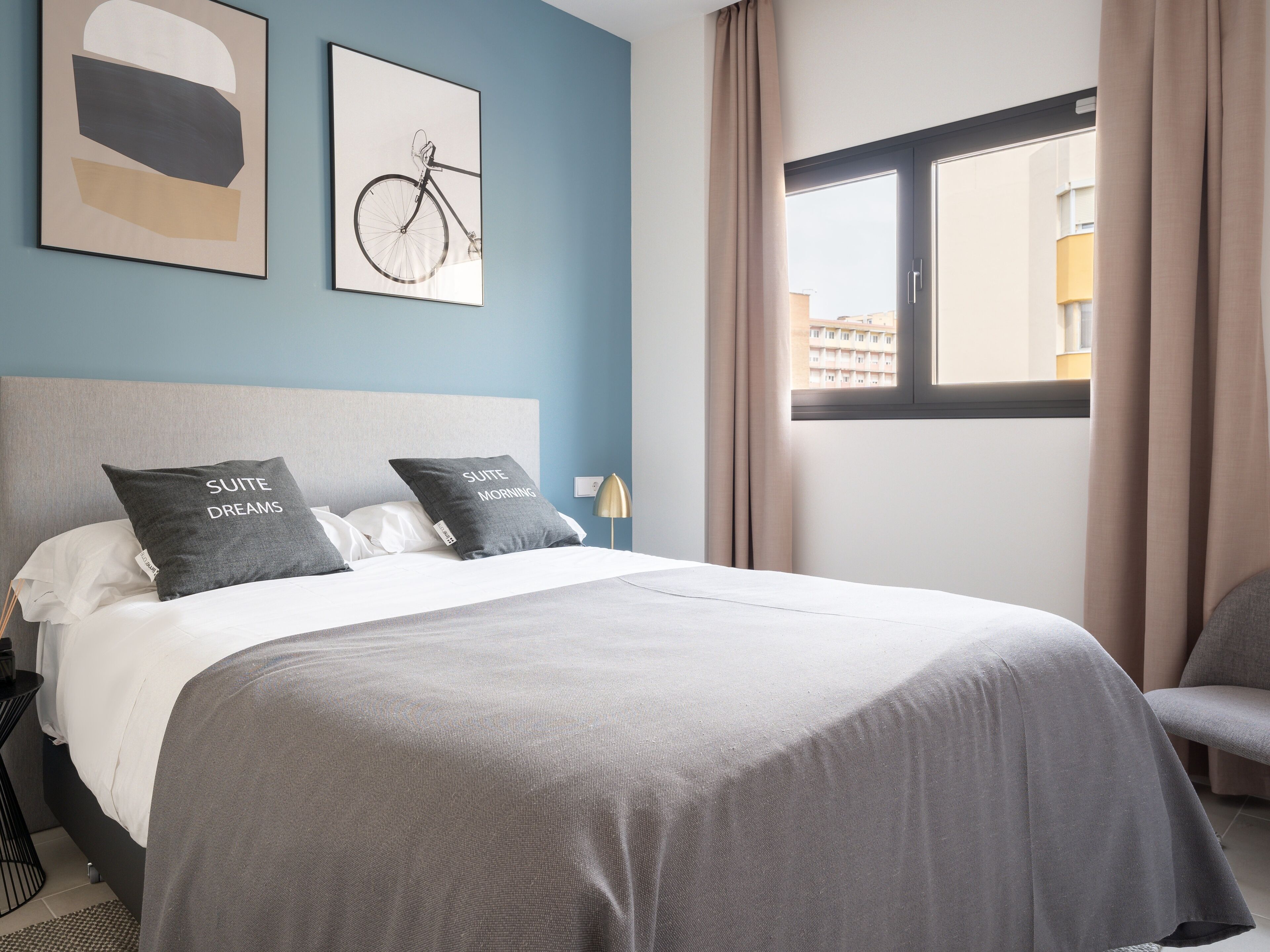 Une chambre d’hôtel avec un grand lit, deux oreillers ornés de motifs, une tête de lit, une fenêtre avec des rideaux et un mur décoré de deux tableaux encadrés.