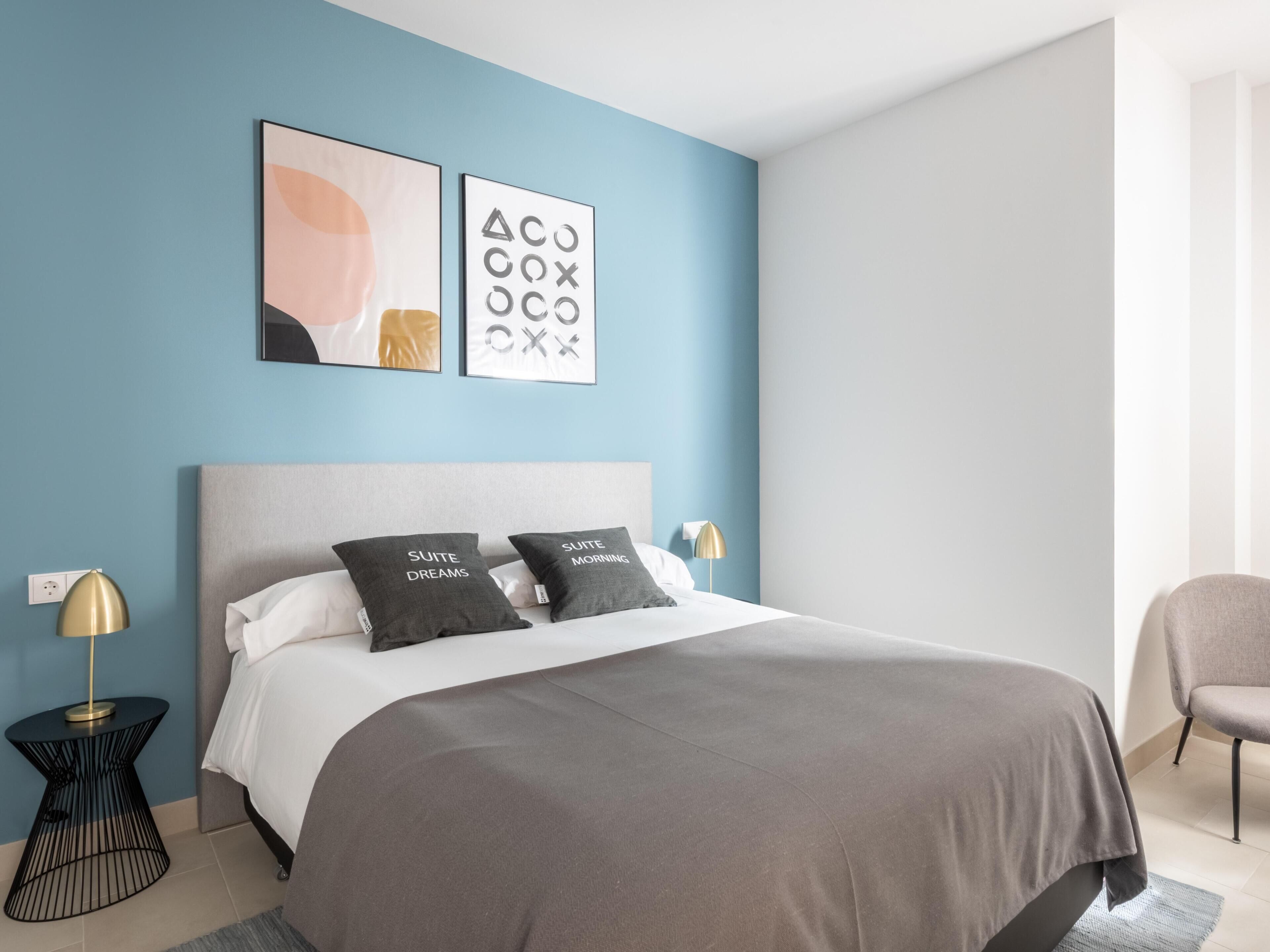 Un dormitorio moderno con una cama gris, dos almohadas con texto, una pared azul y dos mesitas de noche con lámparas.