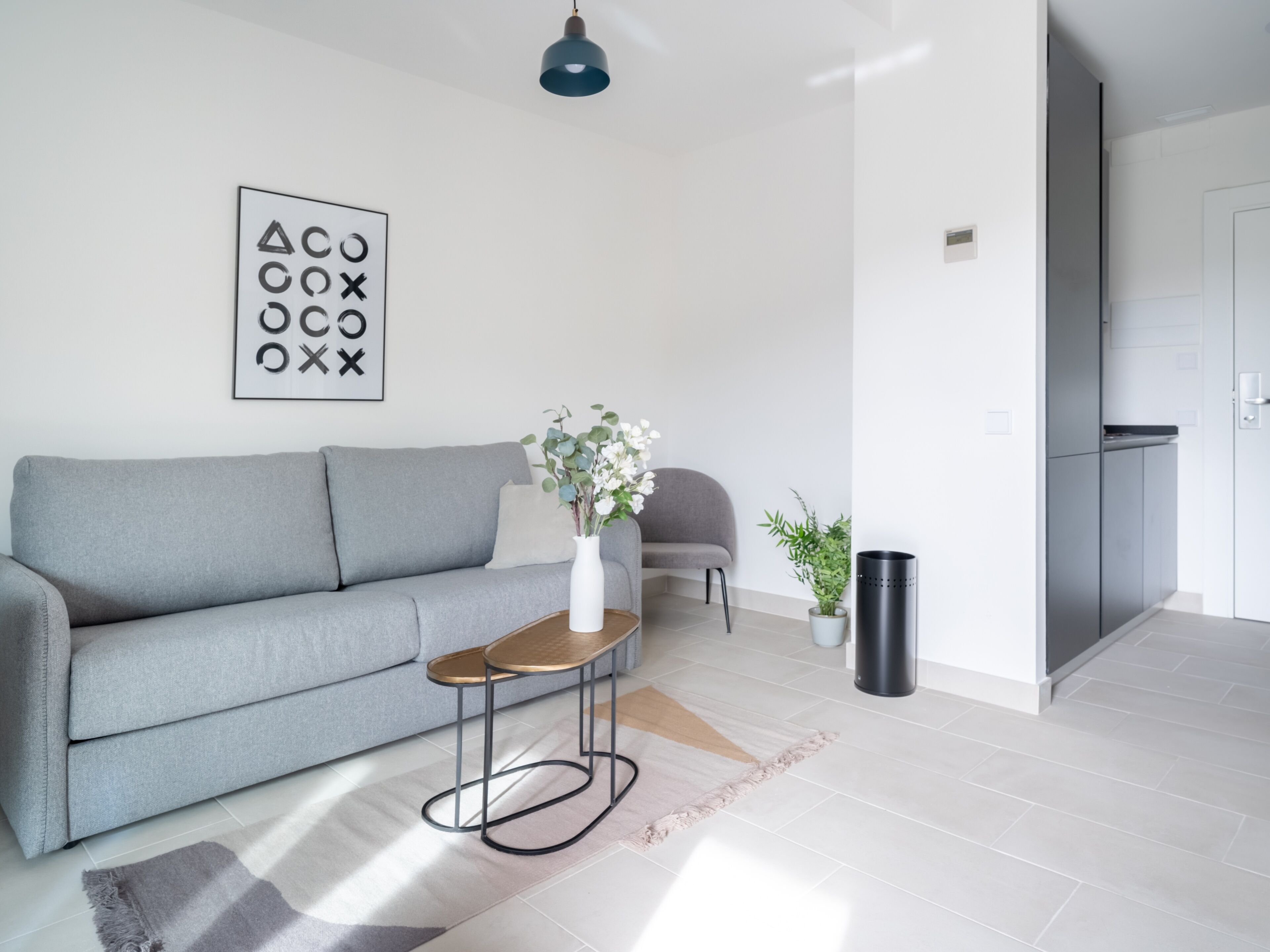Una sala moderna con un sofá gris, una pequeña mesita de madera, un jarrón con flores, un sillón gris, una planta en maceta, un cubo de basura negro y una obra de arte abstracta enmarcada en la pared.