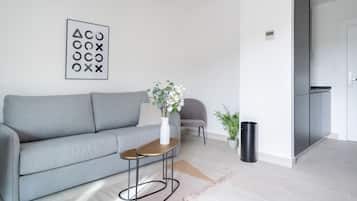 Una sala moderna con un sofá gris, una pequeña mesita de madera, un jarrón con flores, un sillón gris, una planta en maceta, un cubo de basura negro y una obra de arte abstracta enmarcada en la pared.