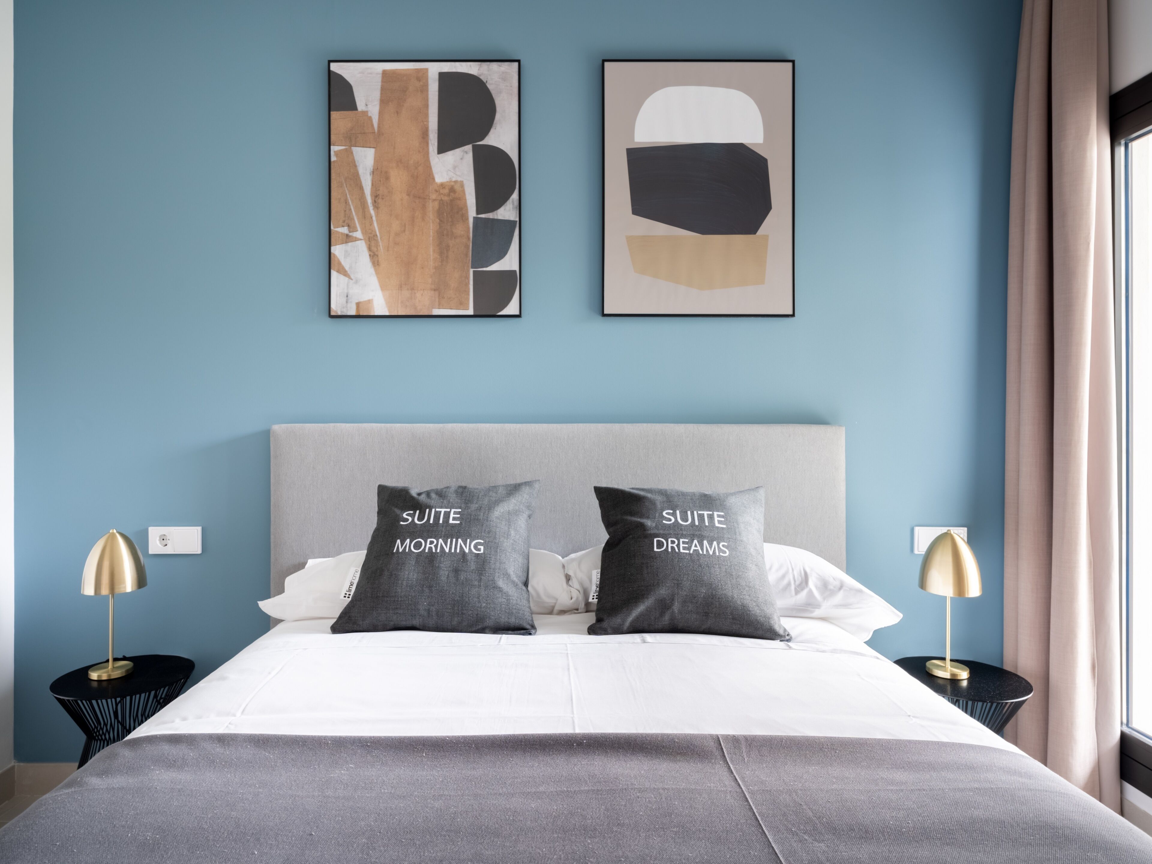 Quarto de hotel com uma cama, duas mesas de cabeceira, um painel de cabeceira e duas pinturas abstratas na parede.