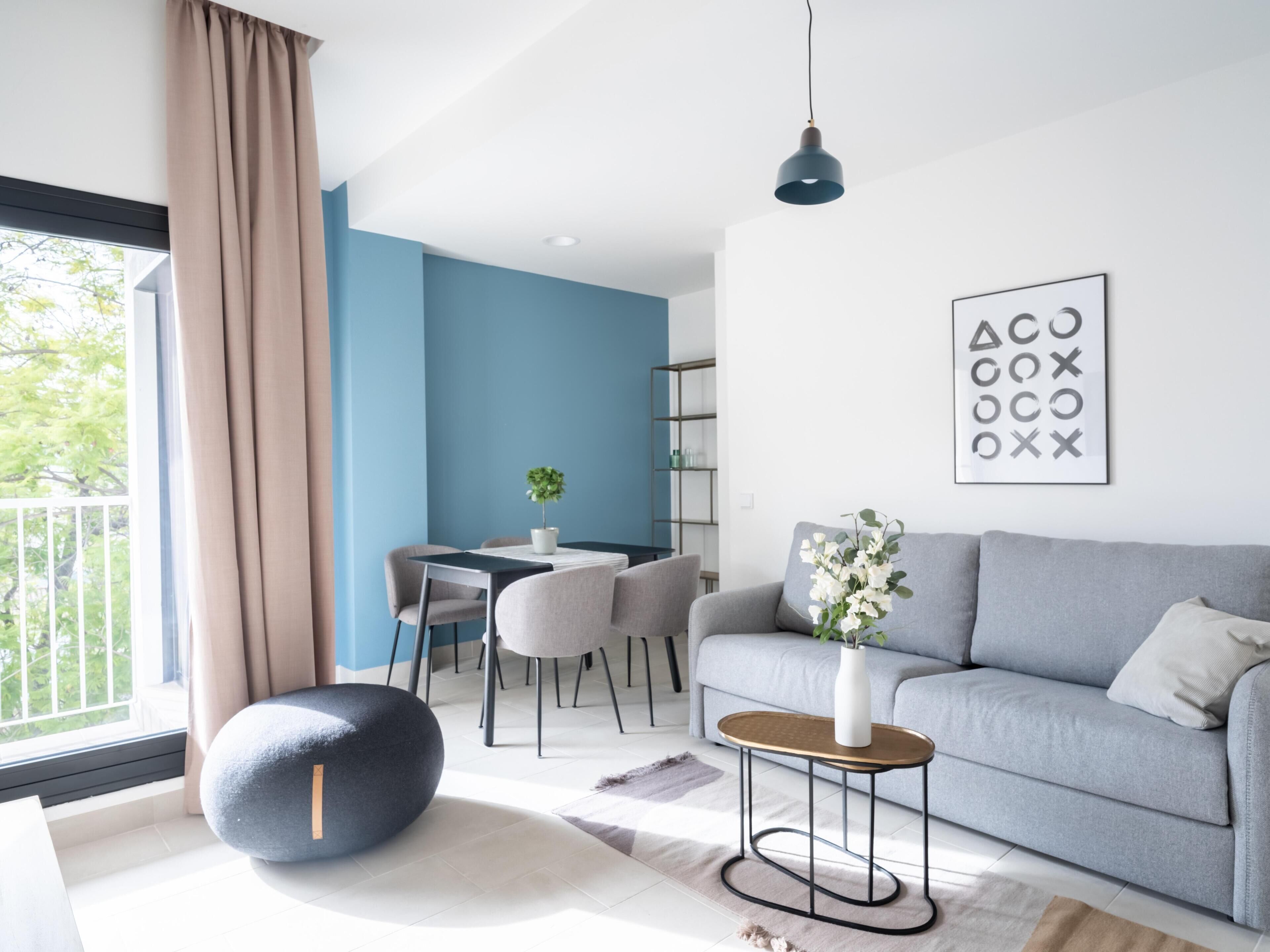 Una sala moderna con un sofá gris, un otoman redondo, una mesita auxiliar y una zona de comedor con mesa y sillas.
