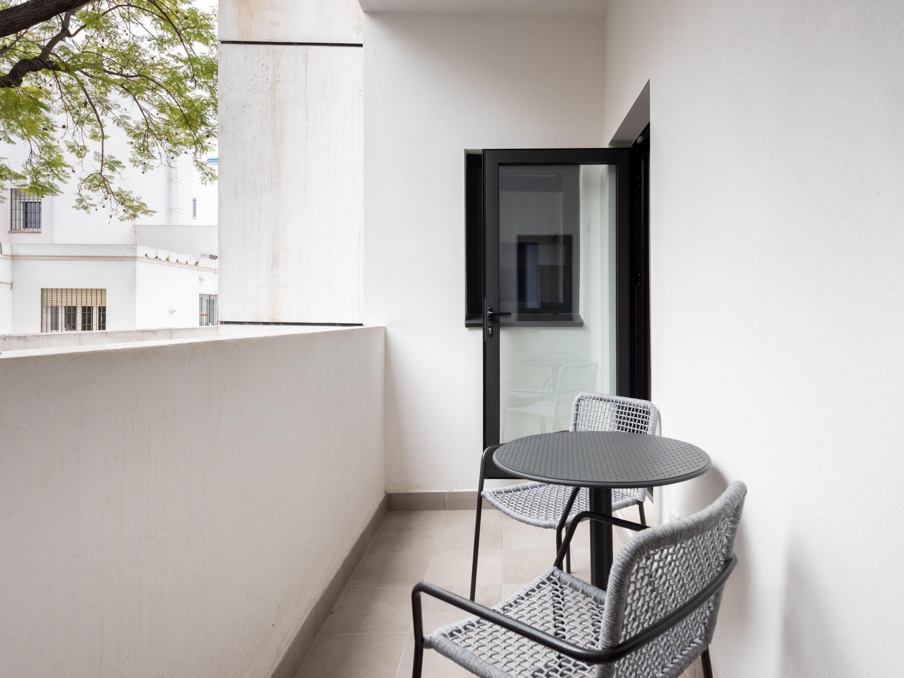 Un balcón con una mesa redonda y dos sillas de mimbre, un vidrio y vistas de edificios al fondo.