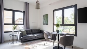 A modern living room with a grey sofa, a round table, and two chairs. There are large windows with curtains, a potted plant, and a flat-screen TV mounted on the wall. - Limehome Sevilla Calle Torcuato Luca de Tena (Seville)