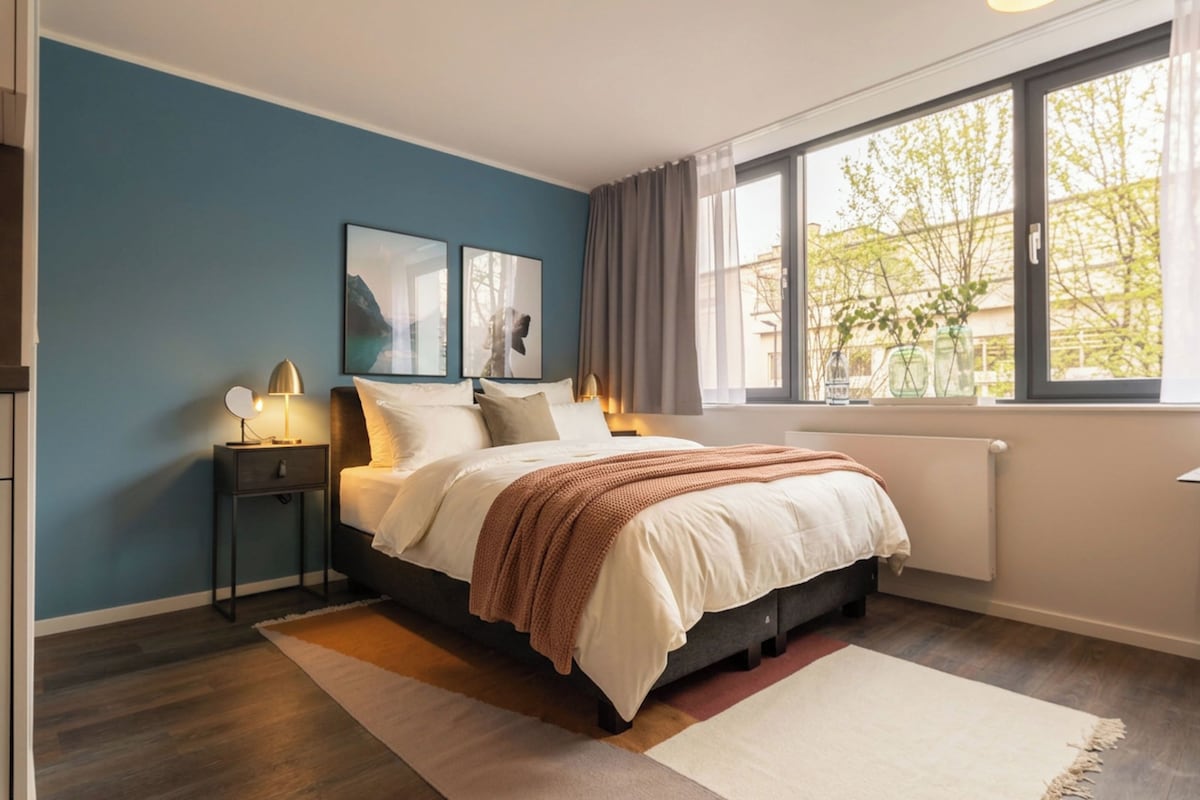 Une chambre à coucher avec un lit, des tables de chevet, une fenêtre avec des rideaux et des œuvres d’art accrochées au mur.