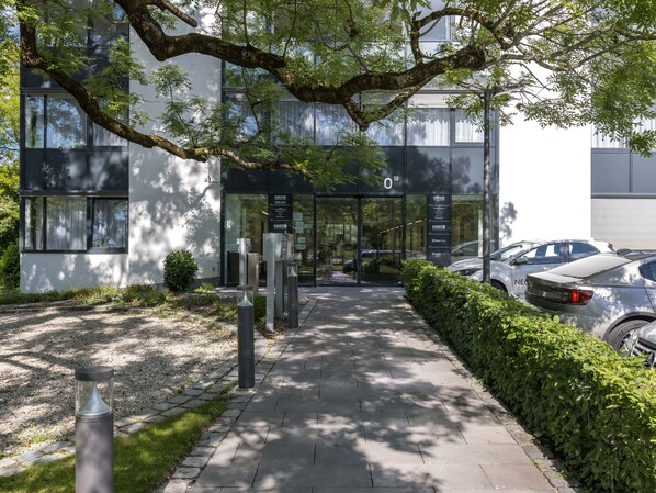 Entrada de un edificio moderno con puerta de vidrio, un cartel y una zona de estacionamiento con coches.