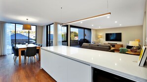 Una sala de estar moderna con un televisor grande, un comedor con mesa y sillas de madera, y una cocina con encimera blanca.