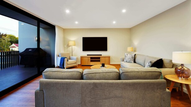 A modern living room with a large grey sofa, a wooden coffee table, a flat-screen TV mounted on the wall, and a balcony with a grill. - Stunning Apartment in the CBD with Parking & WiFi (Launceston)