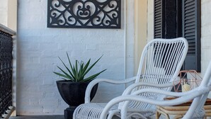 A white wicker chair on a porch with a black metal wall art piece and a potted plant.