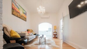 A modern living room with a sofa, leather chairs, a chandelier, a flat-screen TV, and a painting on the wall.