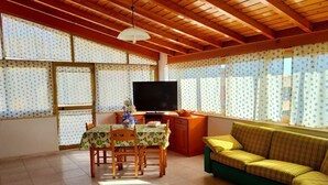Una sala de estar con techo de madera, televisor, sofá, mesa de comedor con sillas y cortinas estampadas.