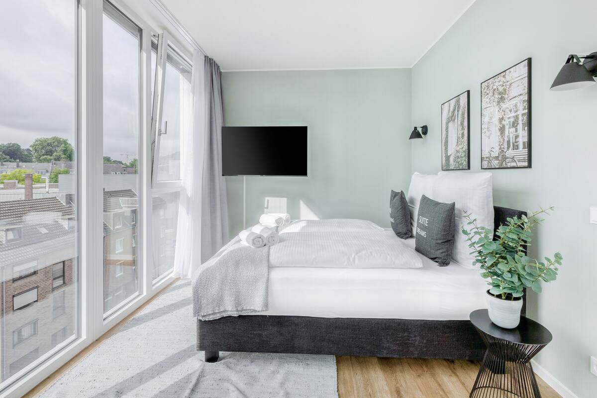Una camera da letto moderna con un'ampia finestra, una televisione a schermo piatto, un letto con cuscini, un comodino con una pianta e opere d'arte appese al muro.