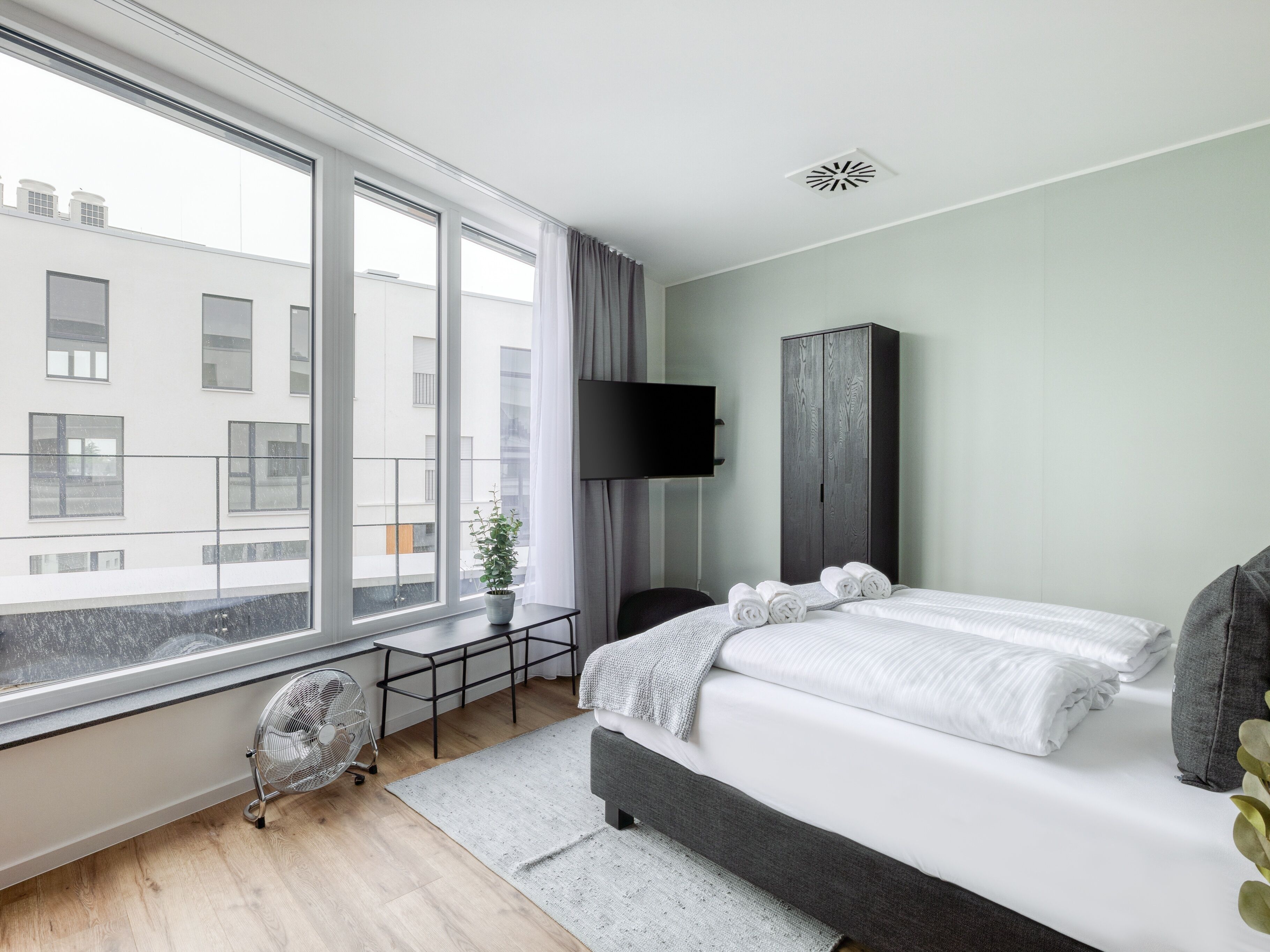 Una camera d'albergo moderna con una grande finestra, un letto con asciugamani ordinatamente piegati, un comodino, una pianta in vaso e un televisore montato a parete.