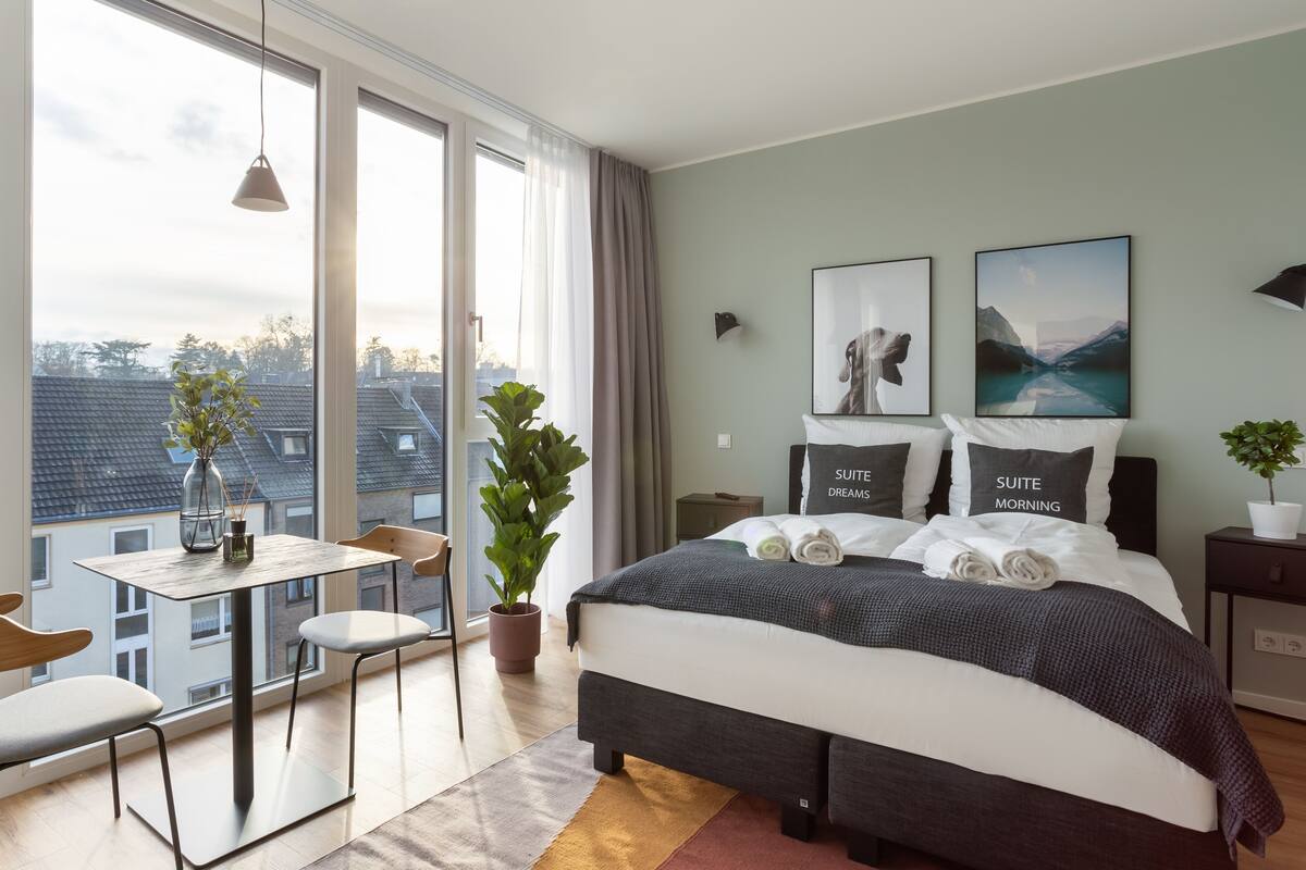 Camera da letto moderna con un letto grande, un tavolino, sedie, una pianta in vaso e vista sugli edifici attraverso ampie finestre.