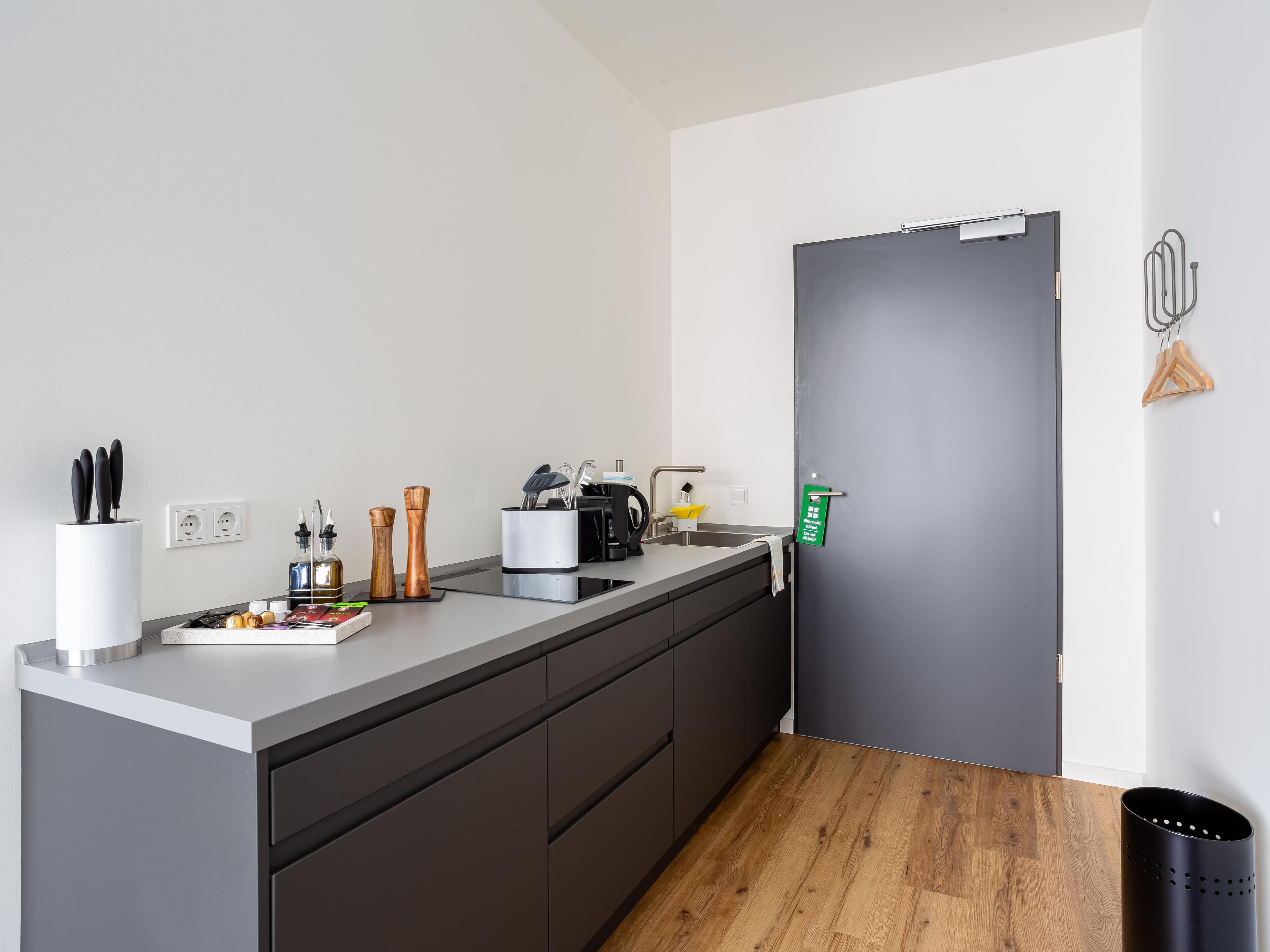 A modern kitchen with a grey countertop, black cabinets, and a wooden floor. There is a coffee machine, a sink, and various kitchen utensils.