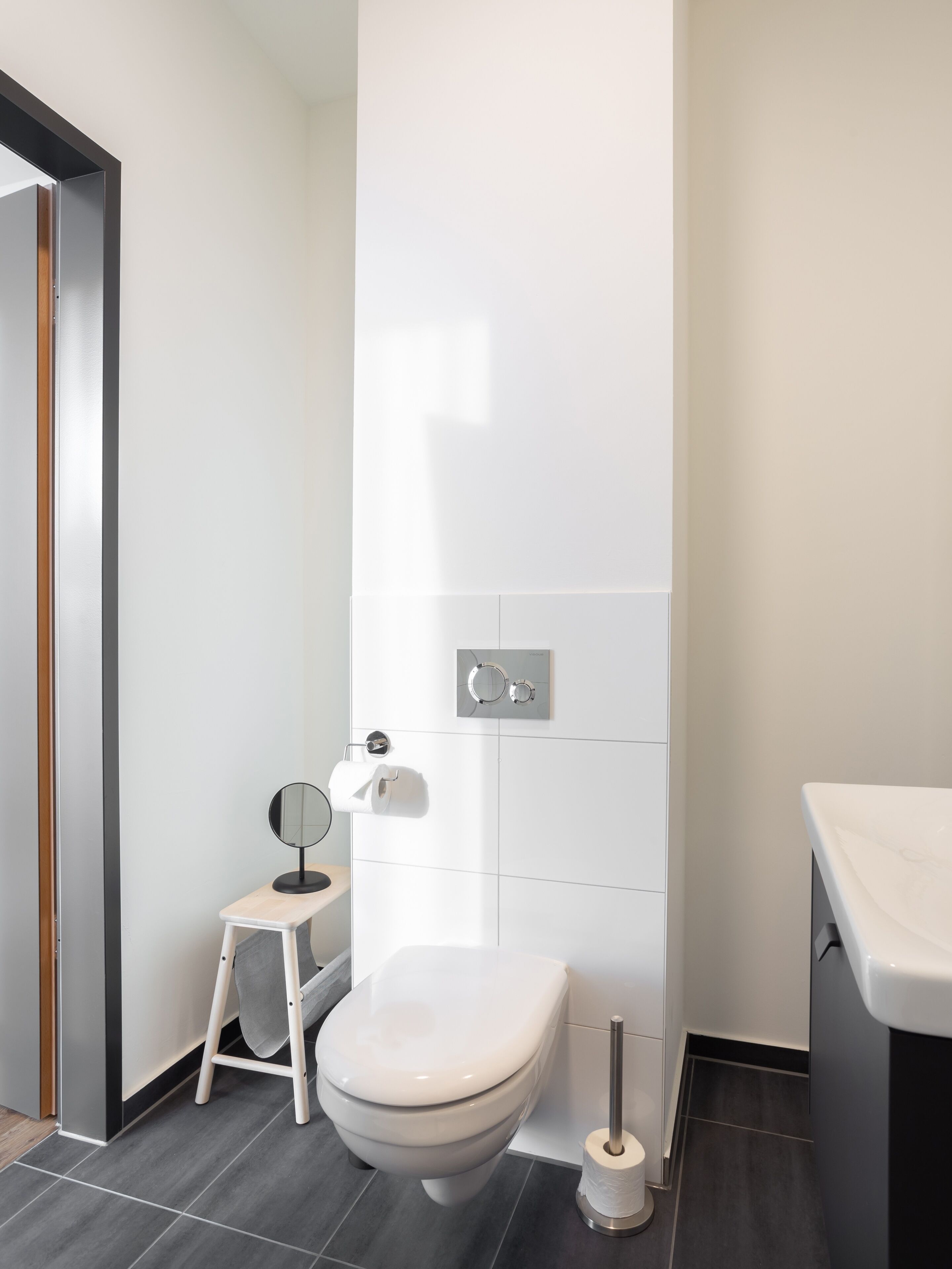 Une salle de bain moderne équipée de toilettes, d’un petit tabouret, d’un miroir et d’un porte-rouleau de papier toilette.